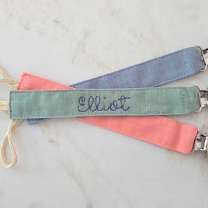 May include: Three baby pacifier clips in pastel colors: pink, blue, and green. The green clip has the name "Elliot" embroidered in blue thread. Each clip has a wooden round end with three holes and a metal clip.