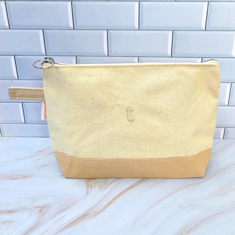 May include: A beige canvas pouch with a tan bottom panel and a zipper closure. The pouch has a small handle on the side and a stitched "t" on the front. The background is a white tile wall.