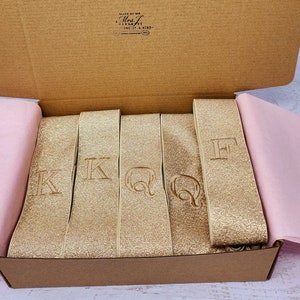 May include: A box filled with gold glitter ribbon with the letters K, Q, and F embroidered on each ribbon.