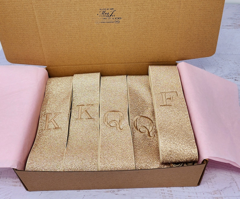 May include: A box filled with gold glitter ribbon with the letters K, Q, and F embroidered on each ribbon.