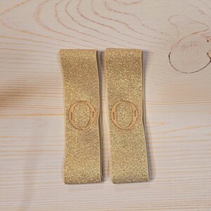 Puede incluir: Dos bandas el&aacute;sticas rectangulares con un acabado de purpurina dorada brillante. Cada banda presenta un parche circular en relieve centrado que contiene la letra O. Las bandas est&aacute;n fabricadas con un material flexible y texturizado de tono dorado.