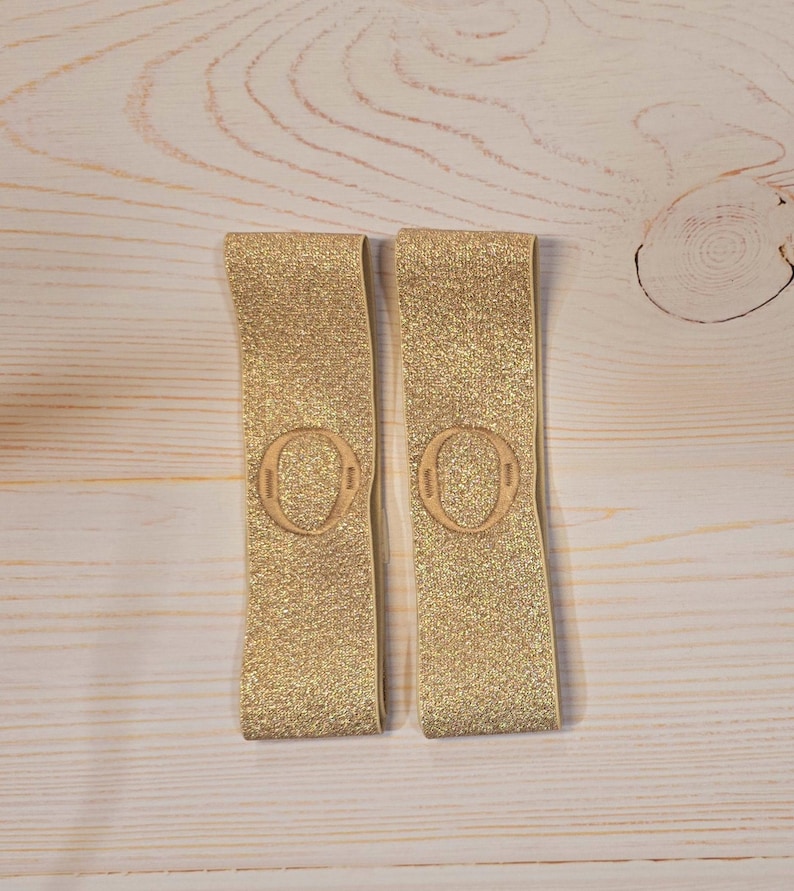 Puede incluir: Dos bandas el&aacute;sticas rectangulares con un acabado de purpurina dorada brillante. Cada banda presenta un parche circular en relieve centrado que contiene la letra O. Las bandas est&aacute;n fabricadas con un material flexible y texturizado de tono dorado.
