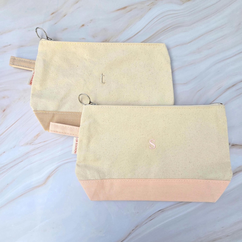 May include: Two canvas cosmetic bags. The bags are a light beige color with a peach-colored bottom. One bag has the letter "t" embroidered on it, and the other has the letter "s". Each bag has a silver zipper and a small loop.