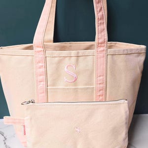 May include: A canvas tote bag and matching pouch set in a light beige color with pink accents. The tote bag and pouch feature an embroidered pink letter "S". The tote bag has pink handles and trim.