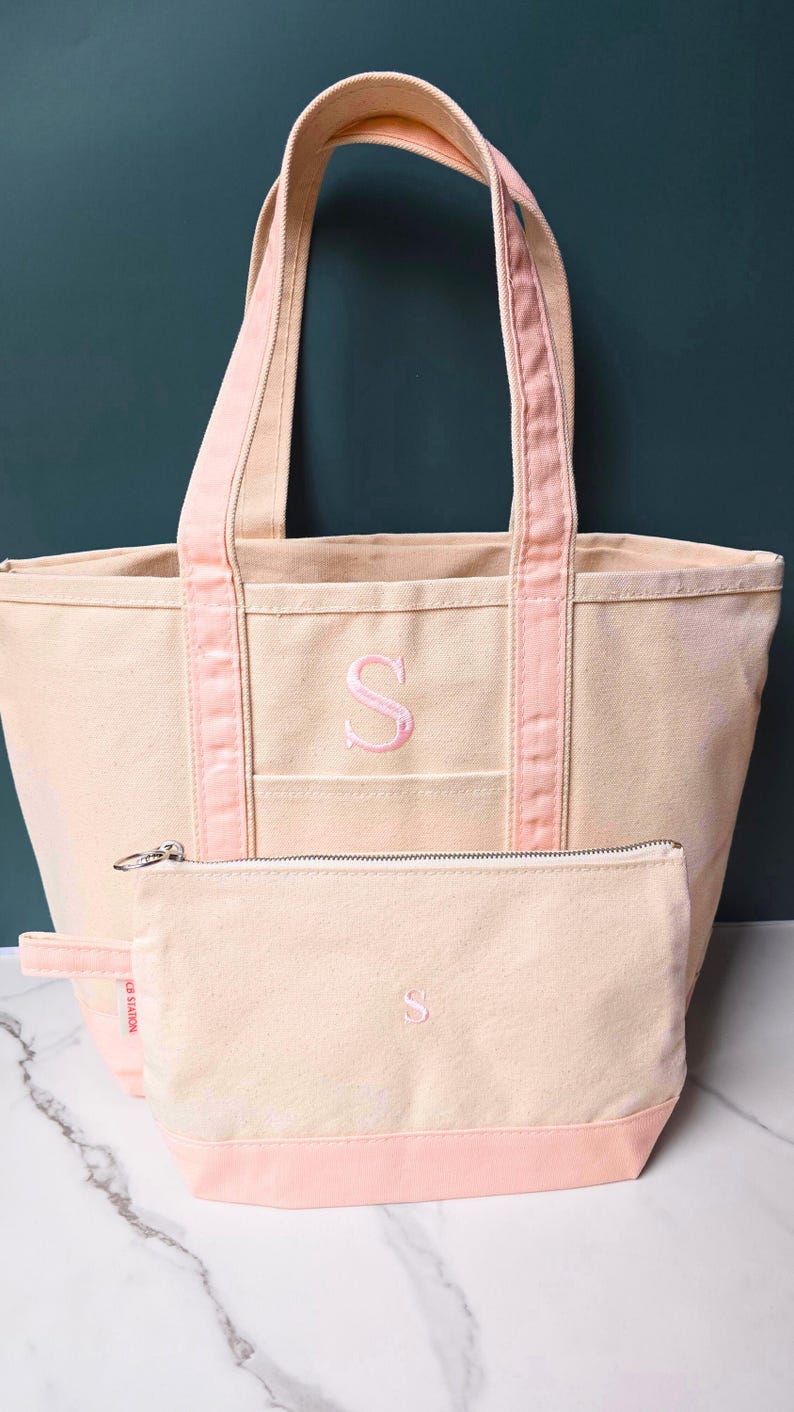 May include: A canvas tote bag and matching pouch set in a light beige color with pink accents. The tote bag and pouch feature an embroidered pink letter "S". The tote bag has pink handles and trim.