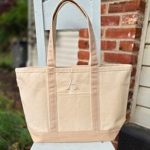 May include: A beige canvas tote bag with a light brown base, straps, and trim. The bag features a large embroidered "A" on the front pocket. The tote is sitting on a light blue chair in front of a brick wall.