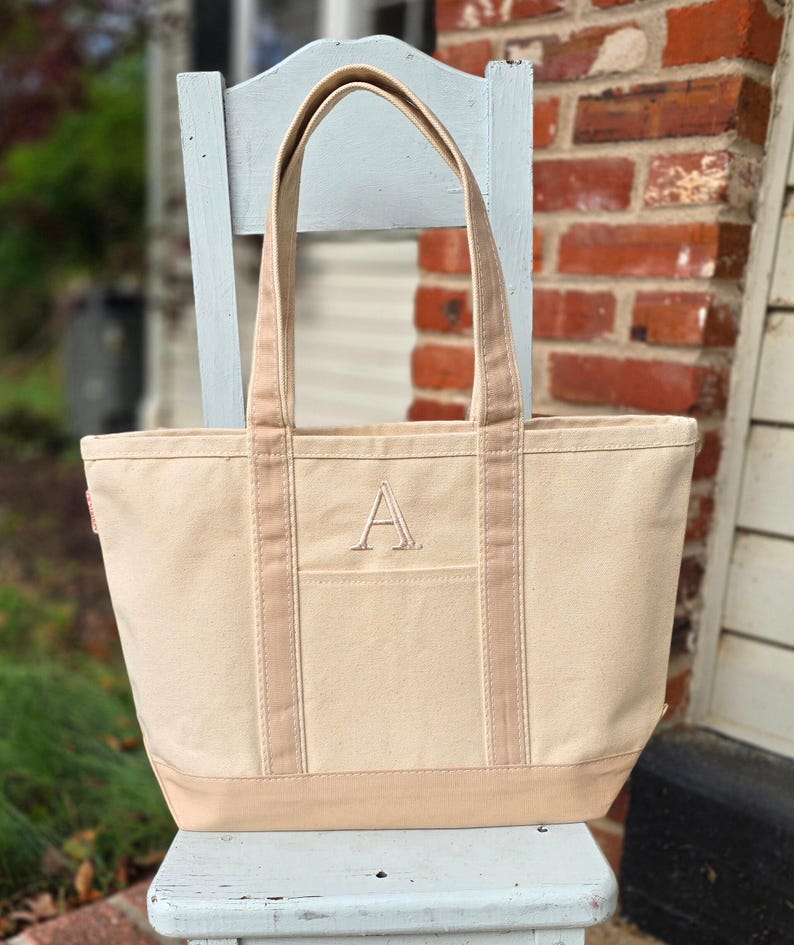 May include: A beige canvas tote bag with a light brown base, straps, and trim. The bag features a large embroidered "A" on the front pocket. The tote is sitting on a light blue chair in front of a brick wall.