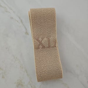 Puede incluir: Una banda el&aacute;stica de color beige doblada en forma rectangular. La tela tiene una apariencia tejida y texturizada, y presenta las letras XL bordadas con hilo beige a juego en el centro de la superficie frontal de la banda.