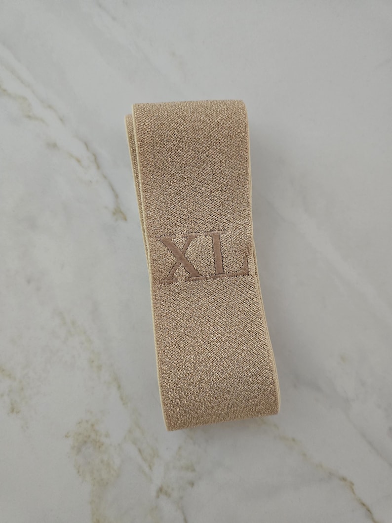 Puede incluir: Una banda el&aacute;stica de color beige doblada en forma rectangular. La tela tiene una apariencia tejida y texturizada, y presenta las letras XL bordadas con hilo beige a juego en el centro de la superficie frontal de la banda.