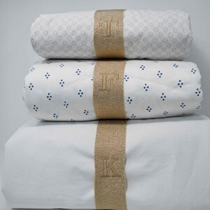 May include: Three white cotton bed sheets with gold trim and embroidered initials "T", "F", and "K".