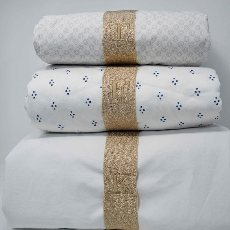 May include: Three white cotton bed sheets with gold trim and embroidered initials "T", "F", and "K".