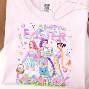 May include: A light pink t-shirt with a graphic featuring the words "Happy Easter" and "Huntrix". The design includes illustrations of anime-style characters wearing bunny ears and holding Easter eggs and baskets. The shirt is made of a soft material.