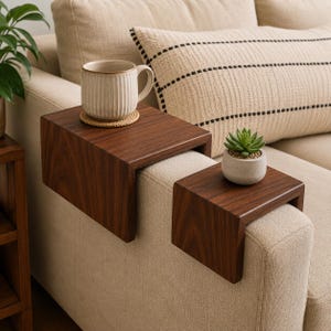 May include: Two dark brown wooden armrest trays with a smooth finish, designed to drape over the arm of a sofa. The larger tray holds a ribbed ceramic mug on a woven coaster, while the smaller tray supports a small potted succulent plant.
