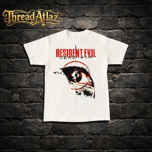 Puede incluir: Camiseta blanca con el texto "RESIDENT EVIL DEAD AIM" en rojo, con un gráfico de un ojo dentro de una retícula. La camiseta tiene mangas cortas y cuello redondo.