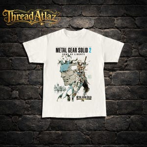 May include: White t-shirt featuring a graphic from the video game Metal Gear Solid 2: Sons of Liberty. The design includes a character portrait and the game's title in black and blue text. The Thread Atlaz logo is at the top.