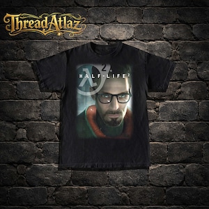 Könnte beinhalten: Schwarzes T-Shirt mit einem Half-Life 2-Grafikdesign, einschließlich des Spiel-Logos und eines Porträts eines Mannes mit Brille. Das Thread Atlaz-Logo ist oben in Gold gehalten. Ein klassisches T-Shirt für Gaming-Fans.