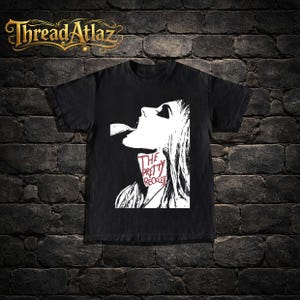 May include: Black t-shirt featuring a white graphic of a woman's profile with the text "THE PRETTY RECKLESS" in red and white. The shirt has a crew neck and short sleeves. The brand logo "Thread Atlaz" is at the top.