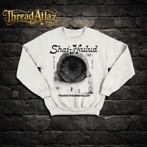 Puede incluir: Sudadera blanca con un gr&aacute;fico en blanco y negro. El dise&ntilde;o incluye el texto "Shai-Hulud" y una ilustraci&oacute;n de una criatura. La frase "FEAR IS THE MIND KILLER" est&aacute; impresa debajo. El logotipo de la marca "Thread Atlaz" est&aacute; en la parte superior.