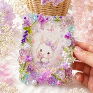 May include: A rectangular picture frame featuring a cartoon bunny illustration. The frame is decorated with colourful beads, flowers, and butterflies in shades of purple, pink, and blue. The bunny holds a bouquet of flowers.