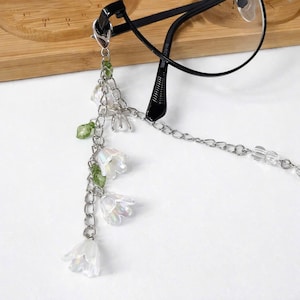 May include: A silver eyeglass chain with a floral design. The chain features small, iridescent white flower charms, green beads, and a delicate chain. The chain is attached to a pair of black spectacles.