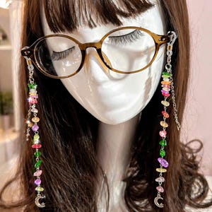 Könnte beinhalten: Braune ovale Brille mit einer dekorativen Kette. Die Kette hat farbige Edelsteinperlen und einen kleinen silbernen Mondsichel-Anhänger. Die Brille wird auf einem Schaufensterkopf mit braunem Haar präsentiert.