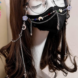 May include: A black face mask and black-rimmed glasses adorned with a silver-toned chain accessory. The chain features small charms including purple flowers, white crescent moons, stars, and colorful beads that drape across the mask and sides.