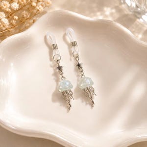 Peut inclure: Une paire d'anneaux de corps non percés argentés avec un design à breloques pendantes. Chaque pièce comprend une petite étoile en métal reliée à un pendentif en forme de méduse translucide et irisée avec des tentacules en métal argenté.