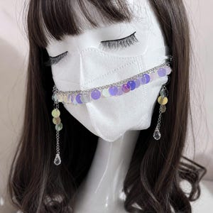 Pode incluir: Uma corrente decorativa para máscara com lantejoulas iridescentes e berloques de cristal. A corrente prateada apresenta uma fileira de lantejoulas redondas e coloridas em tons de roxo, rosa e amarelo. Os berloques de cristal pendem da corrente.