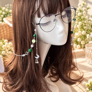 Op de afbeelding: Bril met een zwart montuur en een decoratieve zilverkleurige ketting. De ketting is versierd met groene en lichtgele kralen, heldere kristallen, kleine zilveren sterren en maansikkelbedels die aan de zijkanten van het montuur hangen.
