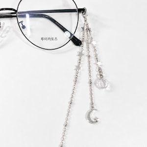 Könnte beinhalten: Runde Brille mit einer silbernen Kette mit Stern- und Mondanhängern. Die Brille hat einen schwarzen Rahmen und klare Gläser. Der Text "루이까또즈" ist auf dem Glas aufgedruckt.