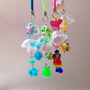 Puede incluir: Una colección de coloridos colgantes de cuentas con varios diseños, incluyendo medusas, peces y flores. Los colgantes están unidos a cordones trenzados en azul, verde y rosa. Algunos colgantes tienen la palabra "YES".