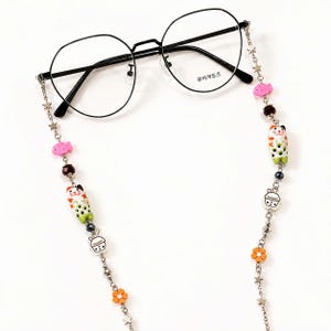 May include: Black round-frame spectacles with a decorative chain. The chain features star, fish, and flower charms, along with a Maneki-neko cat and boba tea cup charm. The text "루이까또즈" is printed on the glasses.