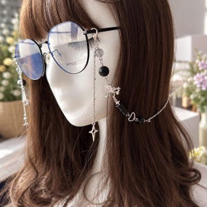 Op de afbeelding: Bril met zwart montuur en een decoratieve zilverkleurige ketting. De ketting is voorzien van zwarte kralen, heldere kristalachtige accenten en kleine zilveren stervormige bedels. De glazen zijn helder en bevatten subtiele tekst op het rechterglas.