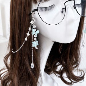 May include: A silver-toned glasses chain featuring dangling light blue flower charms, clear crystal accents, and a delicate star-shaped link chain. The accessory attaches to the frame of black-rimmed glasses with a small lobster claw clasp.