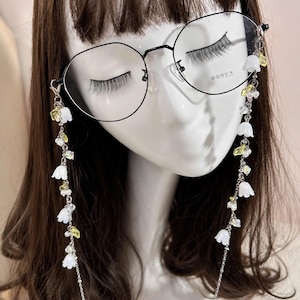 Peut inclure: Lunettes rondes noires avec une chaîne décorative ornée de fleurs blanches et d'accents jaunes. Les lunettes sont présentées sur une tête de mannequin. La chaîne ajoute une touche élégante.