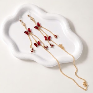 May include: A gold-toned metal chain for eyewear featuring multiple red butterfly-shaped charms with small clear crystals. The chain includes lobster claw clasps at both ends, attached to clear silicone loops for securing to glasses frames.