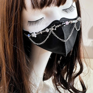 May include: A black face mask featuring a decorative silver-toned chain accessory draped across the front. The chain is adorned with small clear star-shaped charms, iridescent beads, and a black and white patterned bead, creating a layered look.