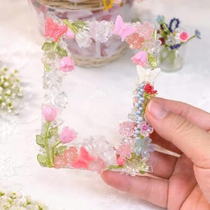May include: A clear, square decorative frame decorated with pink, white, and clear flowers, butterflies, and beads. The frame is held in a hand, with a basket of crocheted flowers in the background. The frame's design includes pink tulips and butterflies.