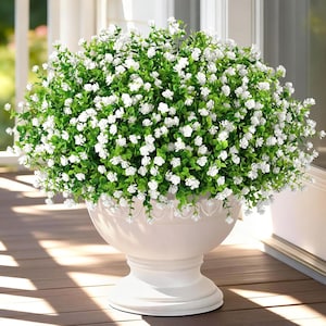May include: A white urn-shaped planter filled with greenery and small white flowers. The planter has a decorative scalloped edge and sits on a pedestal base. The arrangement is a vibrant display of nature.