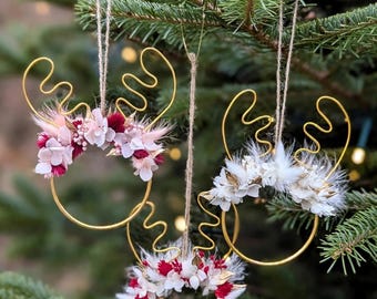 Puede incluir: Tres adornos colgantes con alambre dorado en forma de astas de reno fijados a bases circulares de alambre. Cada adorno está decorado con flores secas en tonos rosa, rojo oscuro y blanco, suspendidos mediante finos lazos de cuerda.