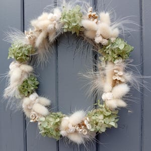 Peut inclure: Une couronne circulaire composée d'hortensias verts séchés, de queues de lapin blanches duveteuses et de délicates plumes blanches. La couronne est posée sur une porte en bois grise vieillie, créant une esthétique rustique et naturelle.