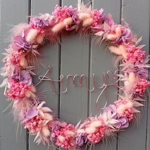 Peut inclure: Une couronne florale circulaire ornée de fleurs roses et violettes, ainsi que d'éléments plumeux. Le nom "Amys" est écrit en fil de cuivre au centre. La couronne est suspendue à une porte en bois grise.
