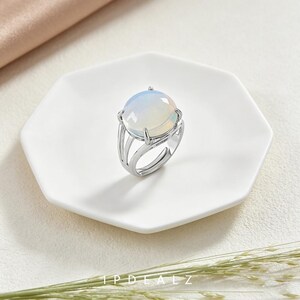 May include: A silver-toned ring featuring a large, round, translucent cabochon stone with an iridescent, milky white appearance. The band is constructed with multiple thin, parallel metal segments that meet at the base of the setting.