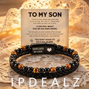 May include: A beaded bracelet featuring alternating black and brown tiger eye beads alongside a braided black band. The clasp is black metal with the text SHIELDED BY LOVE and a heart icon. It is displayed in front of a card with the text TO MY SON.