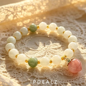 May include: A beaded bracelet featuring smooth, round white and pale green beads separated by small gold-toned spacers. A single peach-shaped pink stone charm with a small green leaf accent hangs from the bracelet, which is marked with the text IPDEALZ.