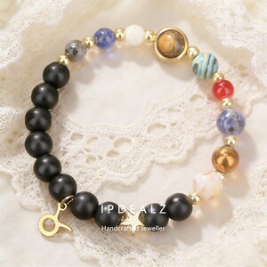 May include: A beaded bracelet featuring black matte beads, gold accents, and colorful gemstone beads. The bracelet includes a gold charm with the Taurus symbol and a star. The text "IPDEALZ Handcrafted Jeweller" is visible.