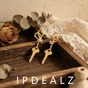 May include: A pair of gold-toned earrings featuring a knotted bow design at the top, accented with small clear stones, and a dangling key-shaped charm at the bottom. The earrings have a matte finish and are secured with a post-back mechanism.