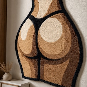 May include: A tufted wall rug featuring a stylized human torso silhouette in shades of tan, beige, and dark brown, outlined with a thick black border. The design depicts a lower back and gluteal area wearing a black thong-style garment.
