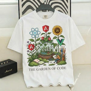 Puede incluir: Camiseta blanca con un diseño de jardín colorido con símbolos de codificación, incluyendo React, Angular, CSS3, Vue y JavaScript. El texto "THE GARDEN OF CODE" está impreso en la parte inferior. La camiseta está colgada en una percha negra.