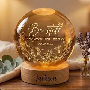 May include: A spherical glass lamp with the text "Be still and know that I am God" and hummingbird and floral designs. The lamp sits on a wooden base engraved with the name "Jackson". Decorative elements include lavender and a succulent.